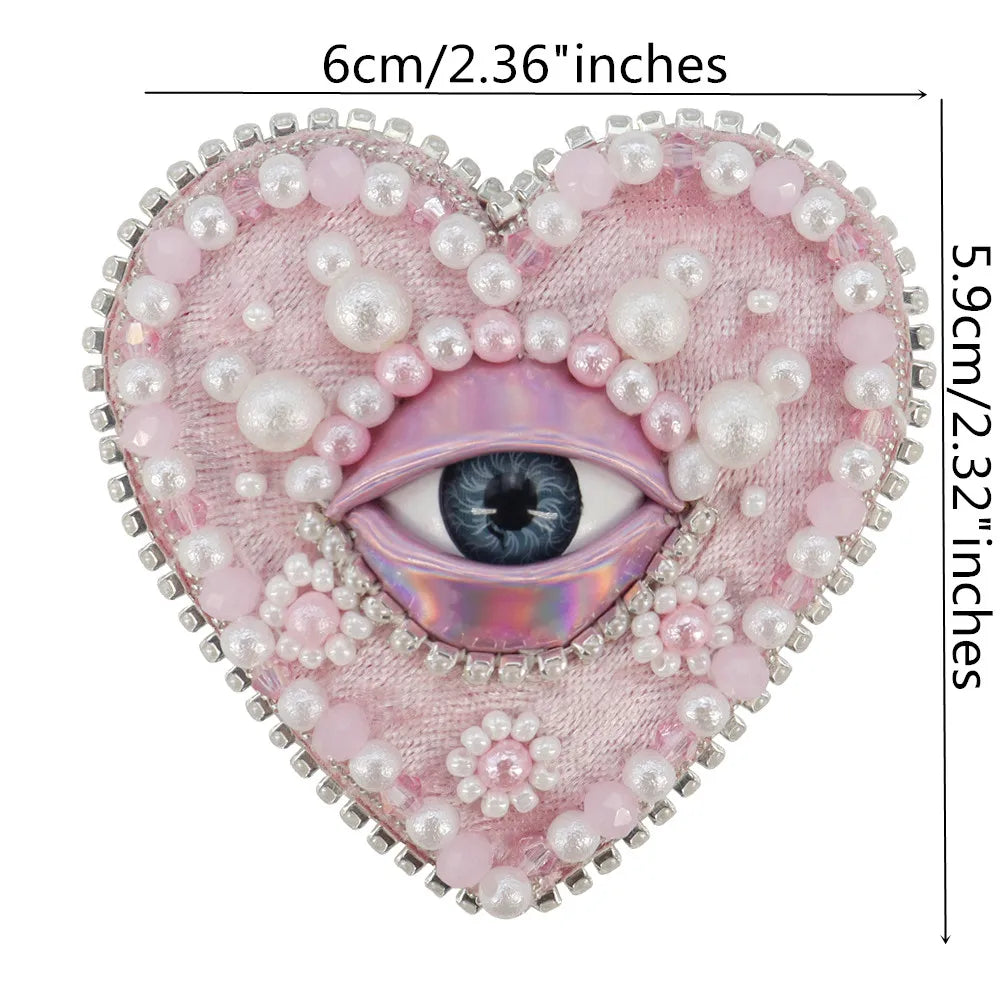 A heart-shaped patch featuring a purple eye surrounded by pink and white beads and rhinestones.