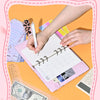 Eine Hand schreibt in einem rosa A6 Budgetplaner-Notizbuch neben Bargeld und einem Taschenrechner.