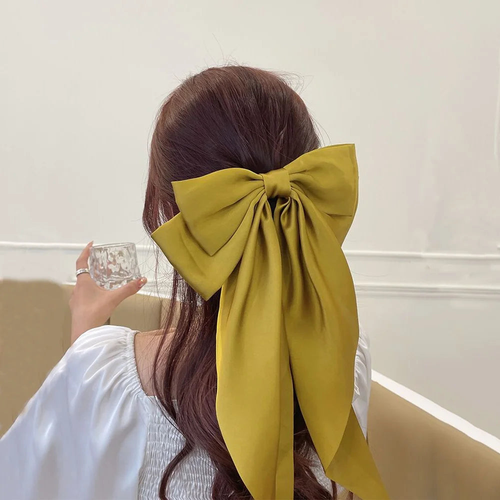 A woman with long brown hair is shown from the back, wearing a white top and a large olive green silk bow tied in her hair.