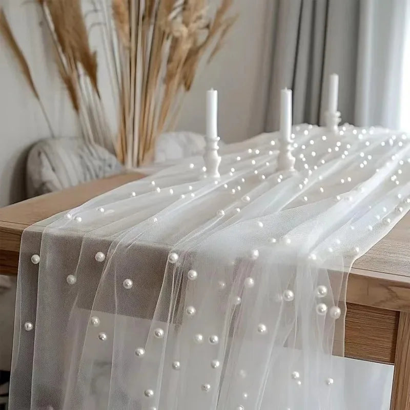 Elegant faux pearl tulle table runner draped over a wooden table with white candles and dried grasses.