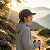 A woman wearing a gray hoodie and a black baseball cap stands outdoors with mountains in the background during sunset.
