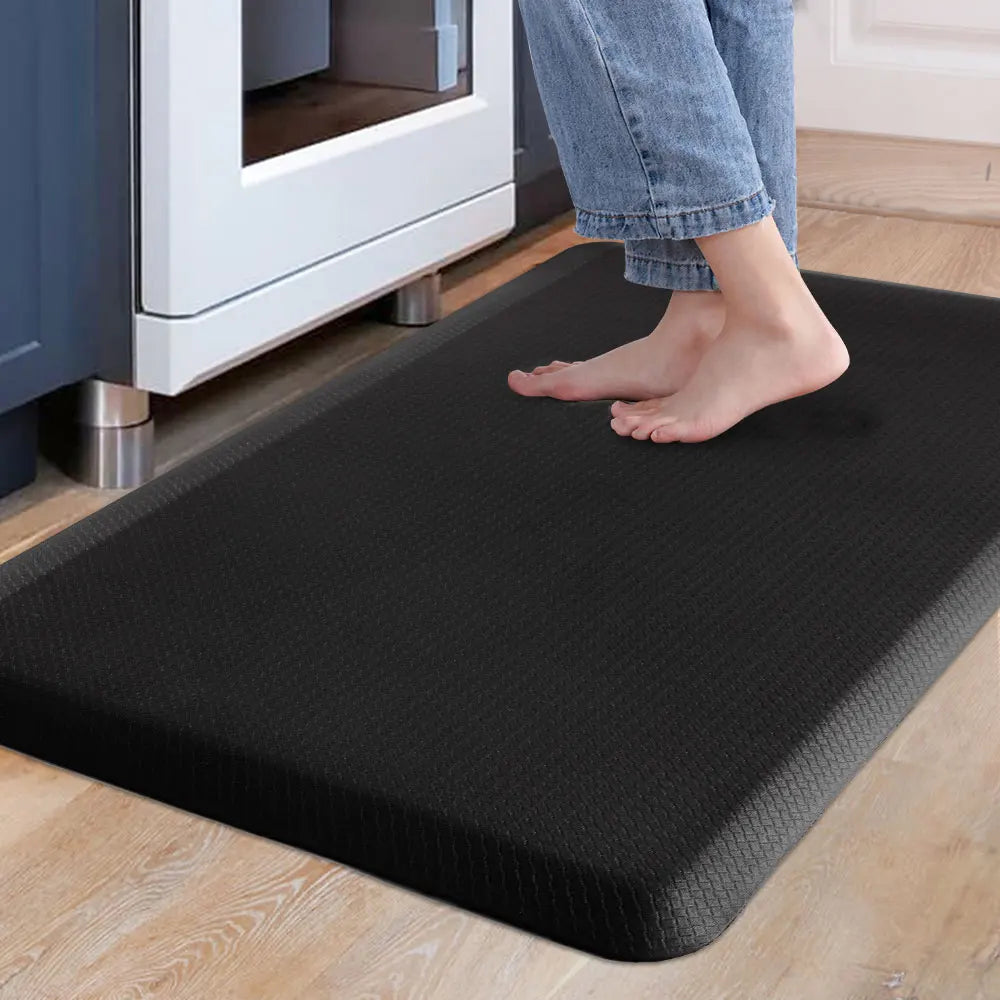A person's feet are standing on a black anti-fatigue mat placed on a light wooden floor near a white appliance.