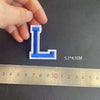 A hand holds a blue embroidered letter L patch, measuring 5.1 by 4.1 cm against a ruler.