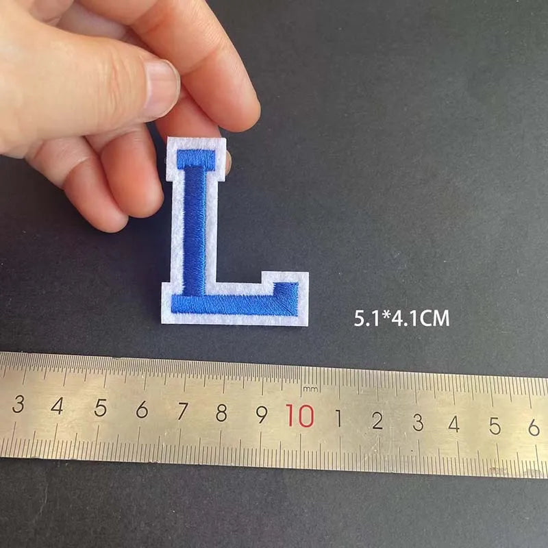 A hand holds a blue embroidered letter L patch, measuring 5.1 by 4.1 cm against a ruler.