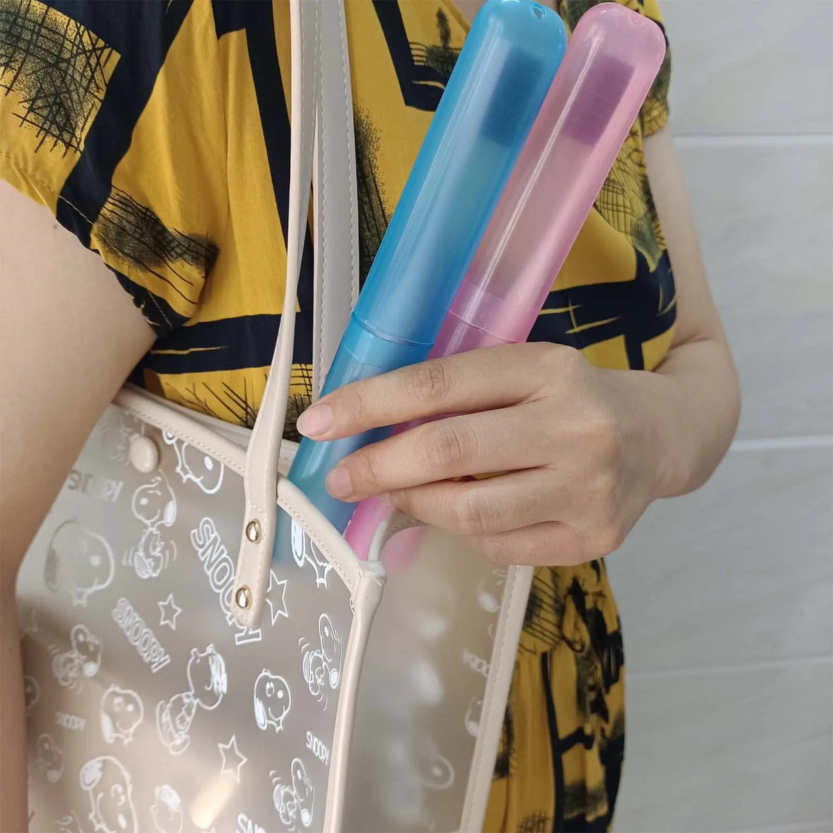 A hand holds a clear silicone travel toothbrush case with pink and blue bristles against a yellow patterned shirt.