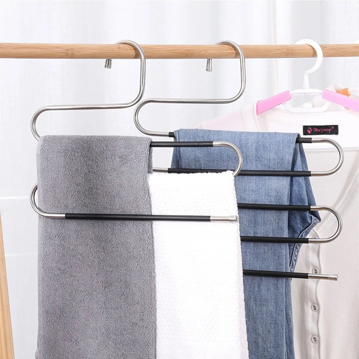 A silver S-shaped pants hanger with multiple bars is shown holding a gray towel, a white towel, and a blue shirt on a rack.