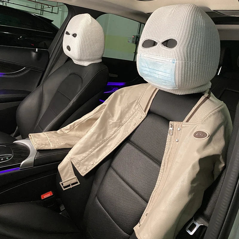 A person wearing a white ski mask head covering and a beige jacket is seated in a car seat.