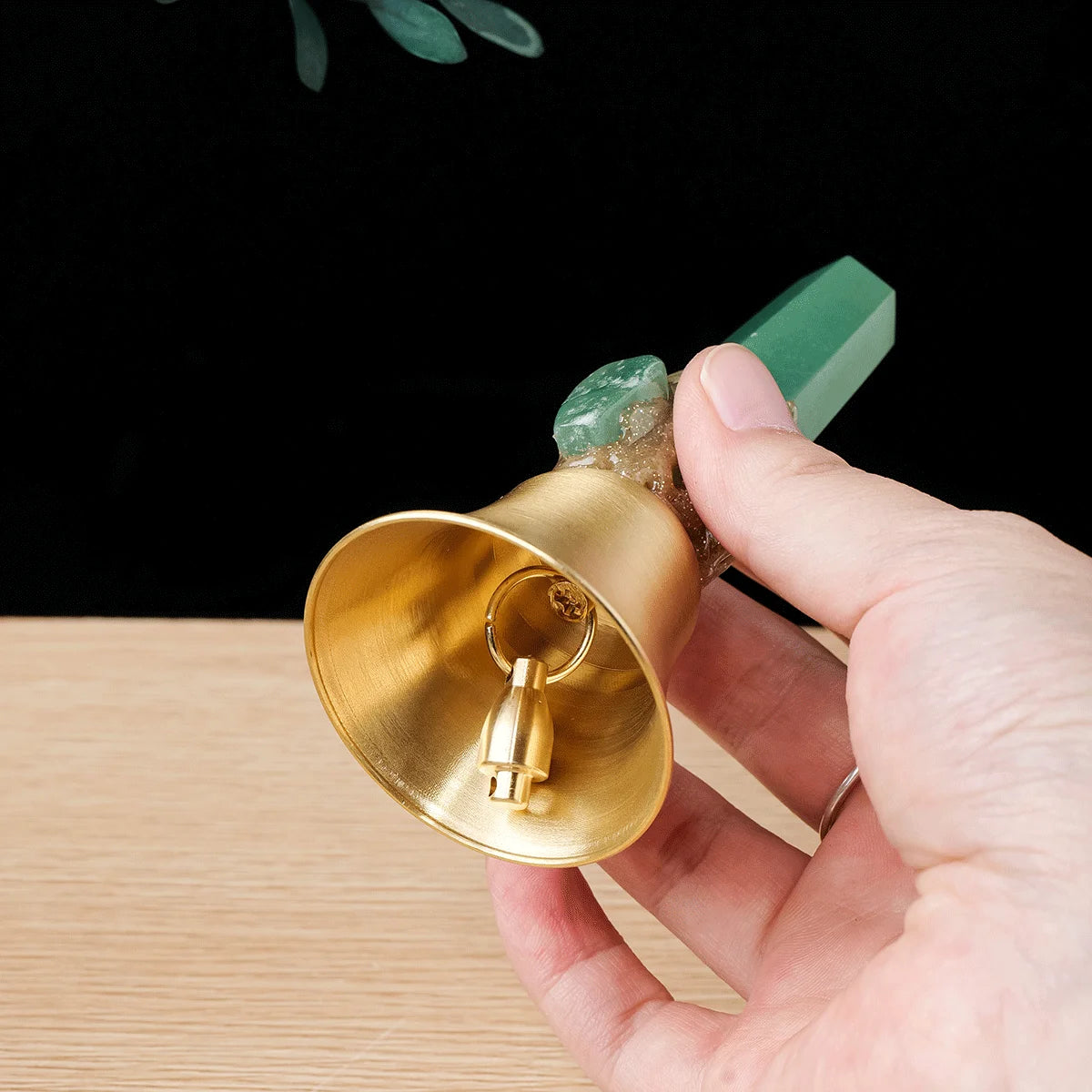 A hand holds a brass bell with a natural green crystal point and a small loop.