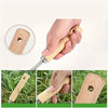 A hand is shown using a stainless steel garden weeder with a wooden handle to remove weeds in grass.
