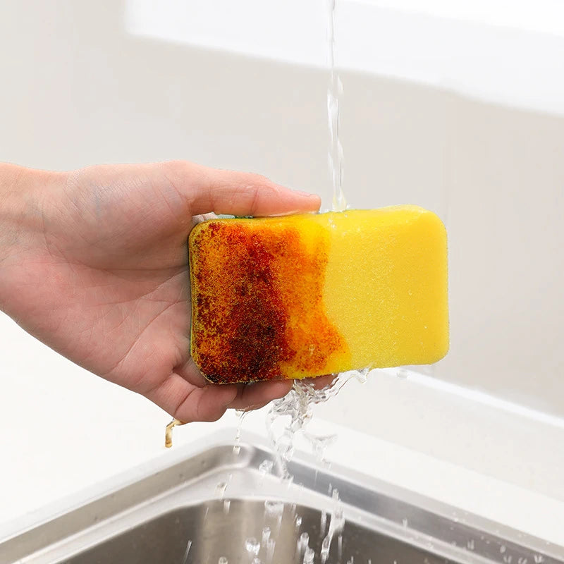 A hand is holding a yellow scouring pad with a dark, used side, as water flows over it in a kitchen sink.