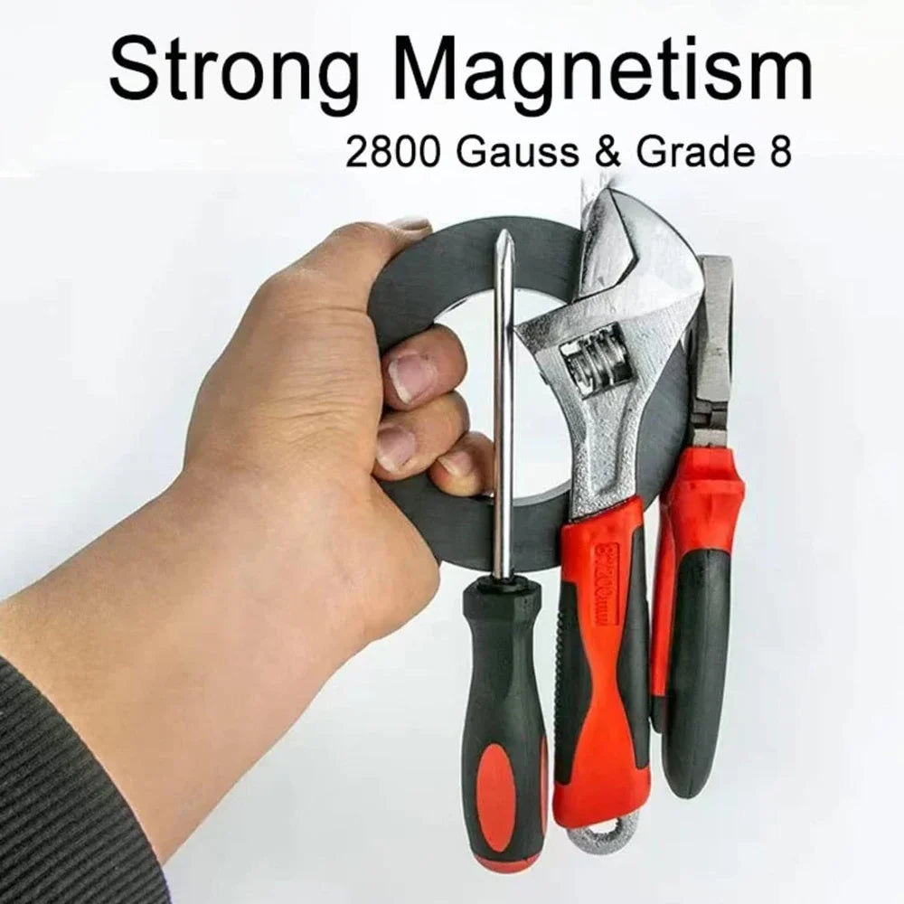 A hand holds a black ceramic ring magnet with a center hole next to various tools, demonstrating strong magnetism.