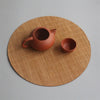 A terracotta teapot and cup resting on a round, woven bamboo tea mat.