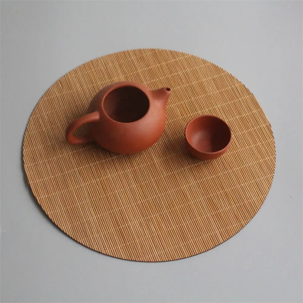 A terracotta teapot and cup resting on a round, woven bamboo tea mat.