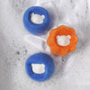 Two round laundry balls, one blue and one orange, each featuring a white cat face, are shown in soapy water.