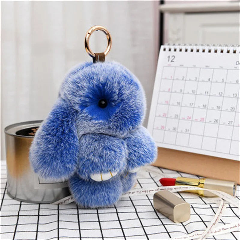 A fluffy blue bunny keychain with a pom pom tail resting on a checkered surface next to a pencil.