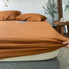 A folded terracotta orange linen fitted sheet resting on a bed, with pillows and a wooden table in the background.