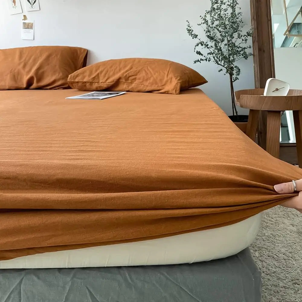 A folded terracotta orange linen fitted sheet resting on a bed, with pillows and a wooden table in the background.