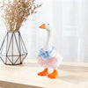 A white ceramic goose figurine wearing a blue floral outfit and orange feet, displayed next to dried flowers.