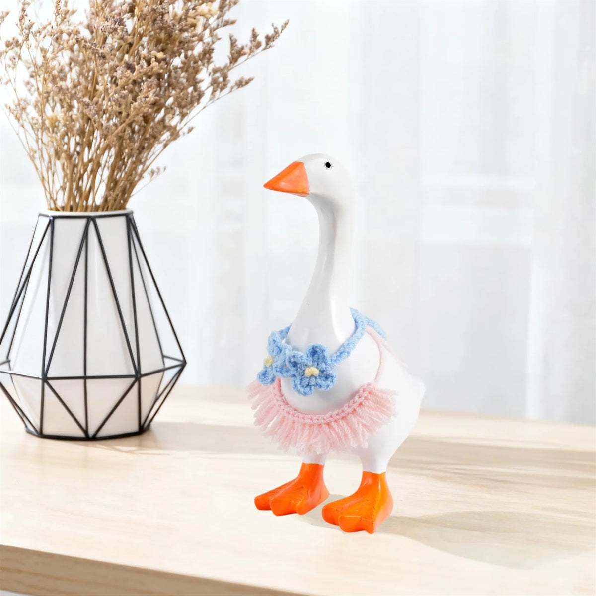 A white ceramic goose figurine wearing a blue floral outfit and orange feet, displayed next to dried flowers.