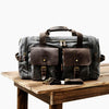 A dark gray canvas duffel bag with brown leather trim and pockets resting on a wooden surface.