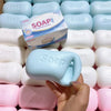 A hand holds a light blue soap-shaped stress relief squishy fidget toy surrounded by other white and pink soaps.