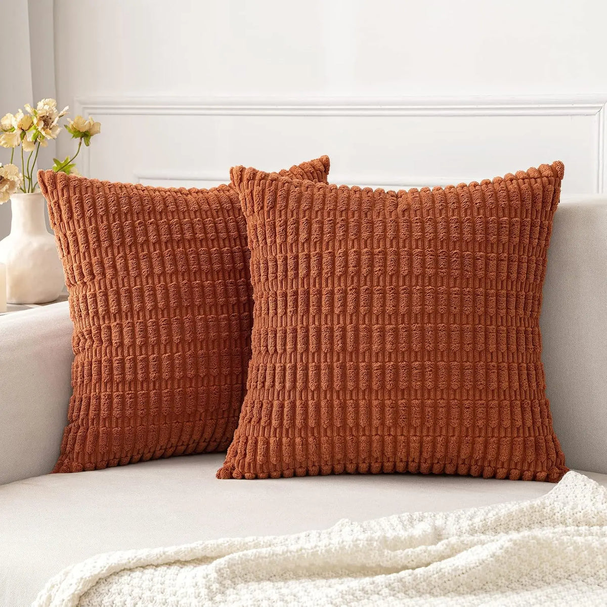 A square, textured rust-colored corduroy throw pillow resting on a light sofa next to a white blanket.
