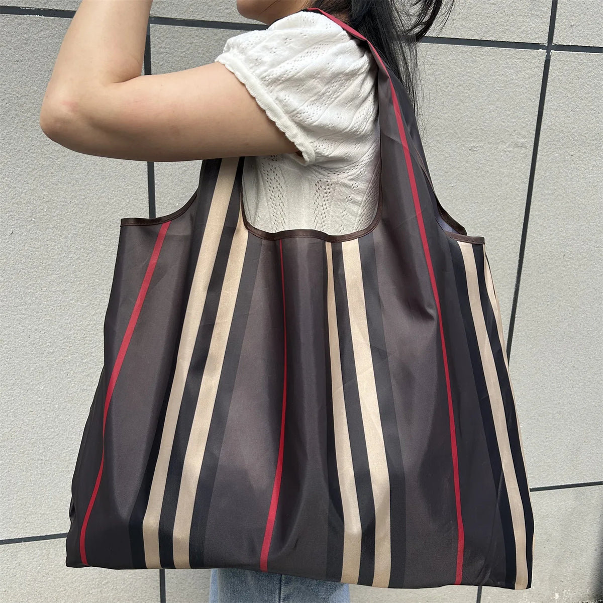 A person is holding a brown and beige striped foldable oxford cloth tote bag against a light gray wall.