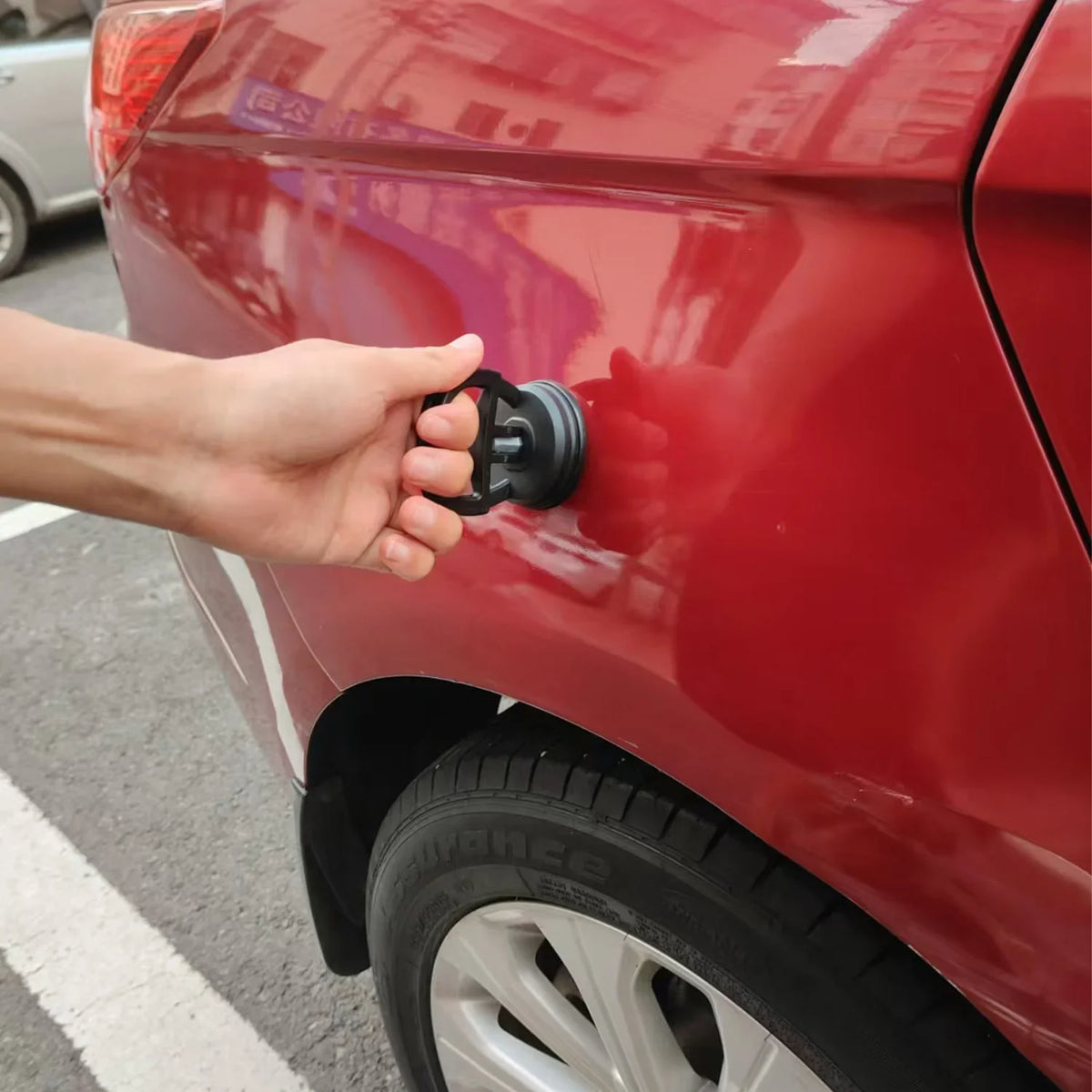 A hand is operating a car dent puller kit with a handle lift and power remover on the rear fender of a red car.