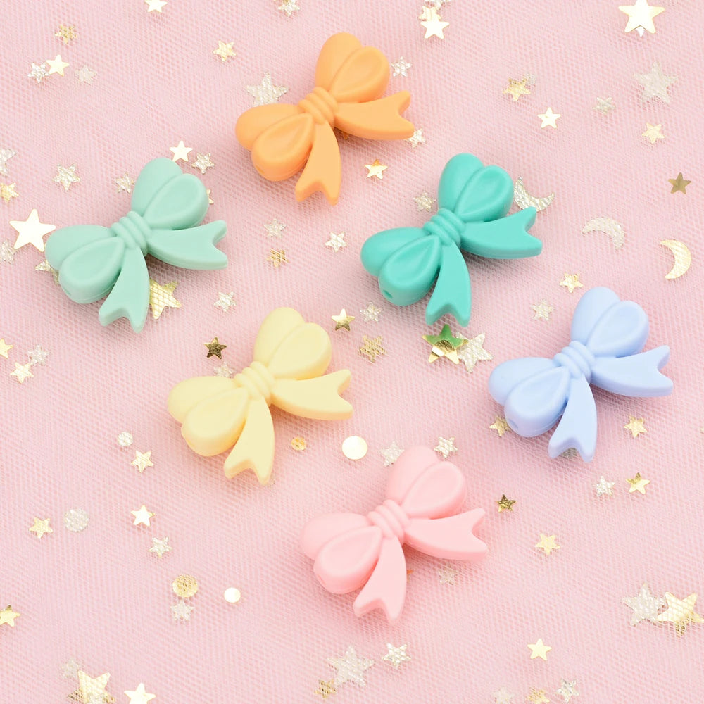 A collection of colorful silicone bow beads in peach, orange, mint green, yellow, blue, and pink scattered on a pink surface 