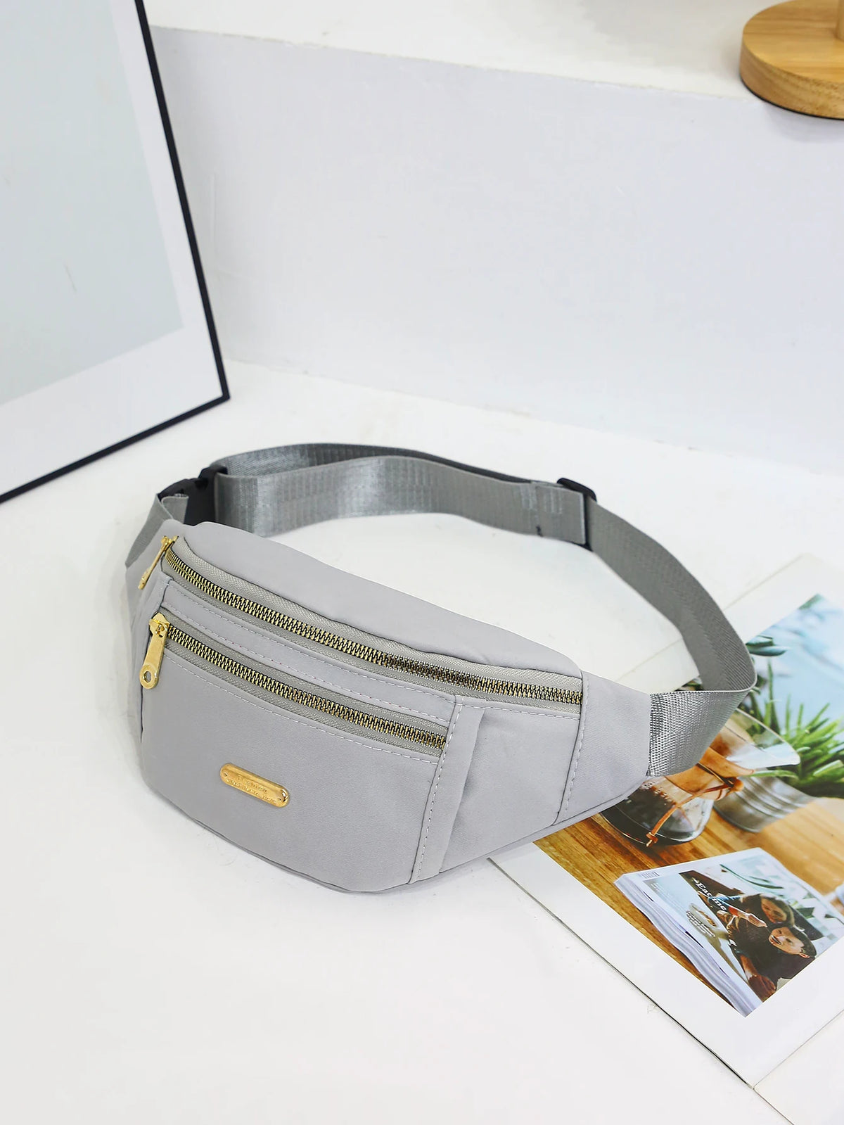 A light gray nylon waist pack with a zipper and gold accent is displayed on a white surface.