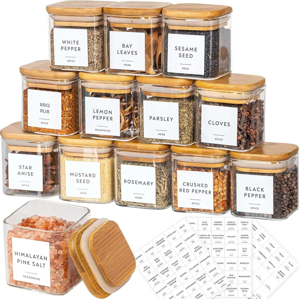 A collection of five square glass spice jars with bamboo lids and labels, including white pepper, bay leaves, sesame seed, le