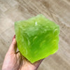 A hand holds a translucent lime green frosted matte jumbo cheese stress ball against a blurred background.