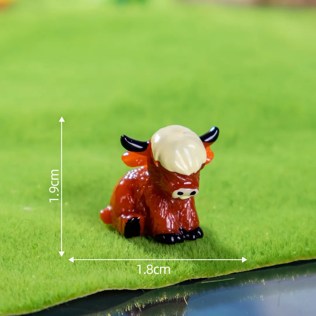 A small, reddish-brown resin figurine of a Highland cow with horns and white fur, resting on green grass.