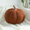 A round, textured brown pumpkin plush pillow with a fuzzy stem resting on a white knitted fabric.