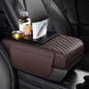 A dark brown quilted car armrest cushion with storage and cup holders is placed on a black car seat.