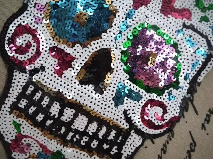 A close-up of a colorful sequin skull embroidered patch featuring purple, teal, and pink hues.