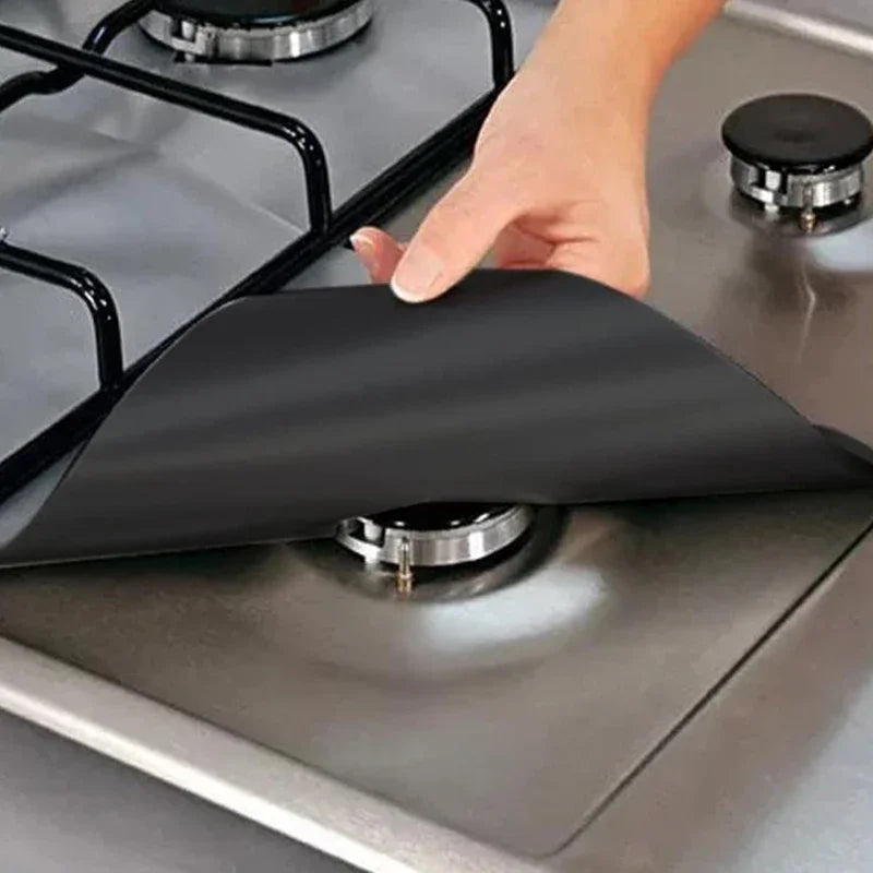 A hand places a black square gas hob protector liner onto a stainless steel stovetop.