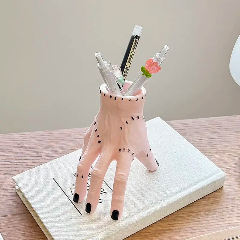 A pink resin Halloween hand planter and pen holder with black dots, holding several pens and a small flower.