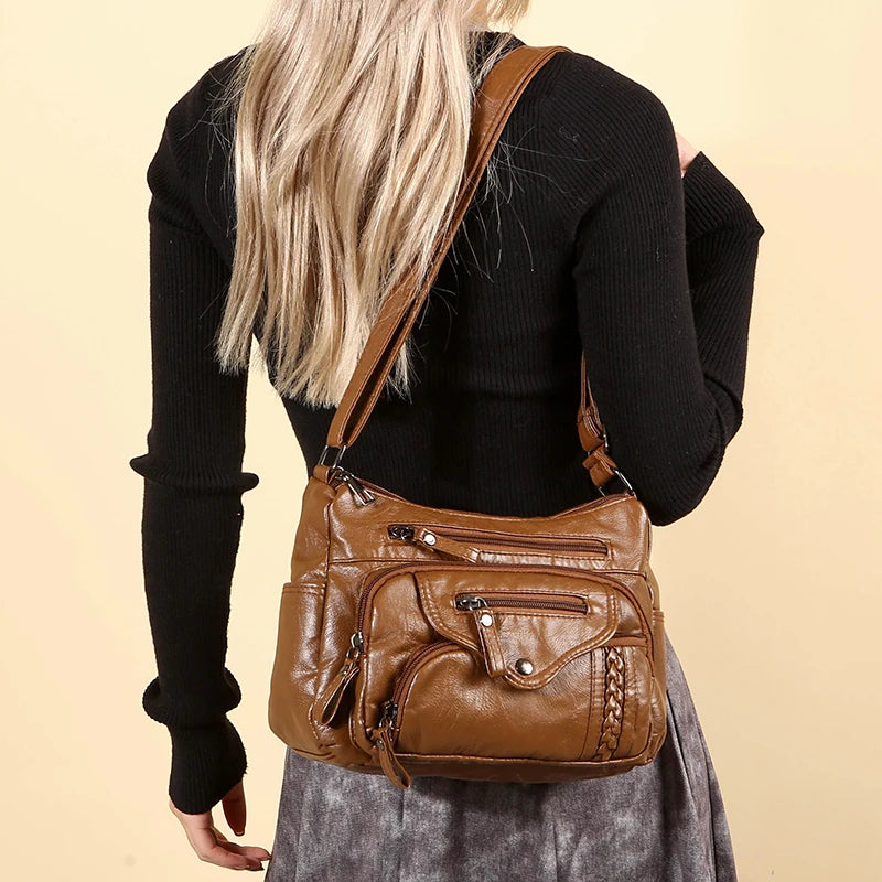 A woman is holding a brown leather tote bag with an adjustable strap against a light background.