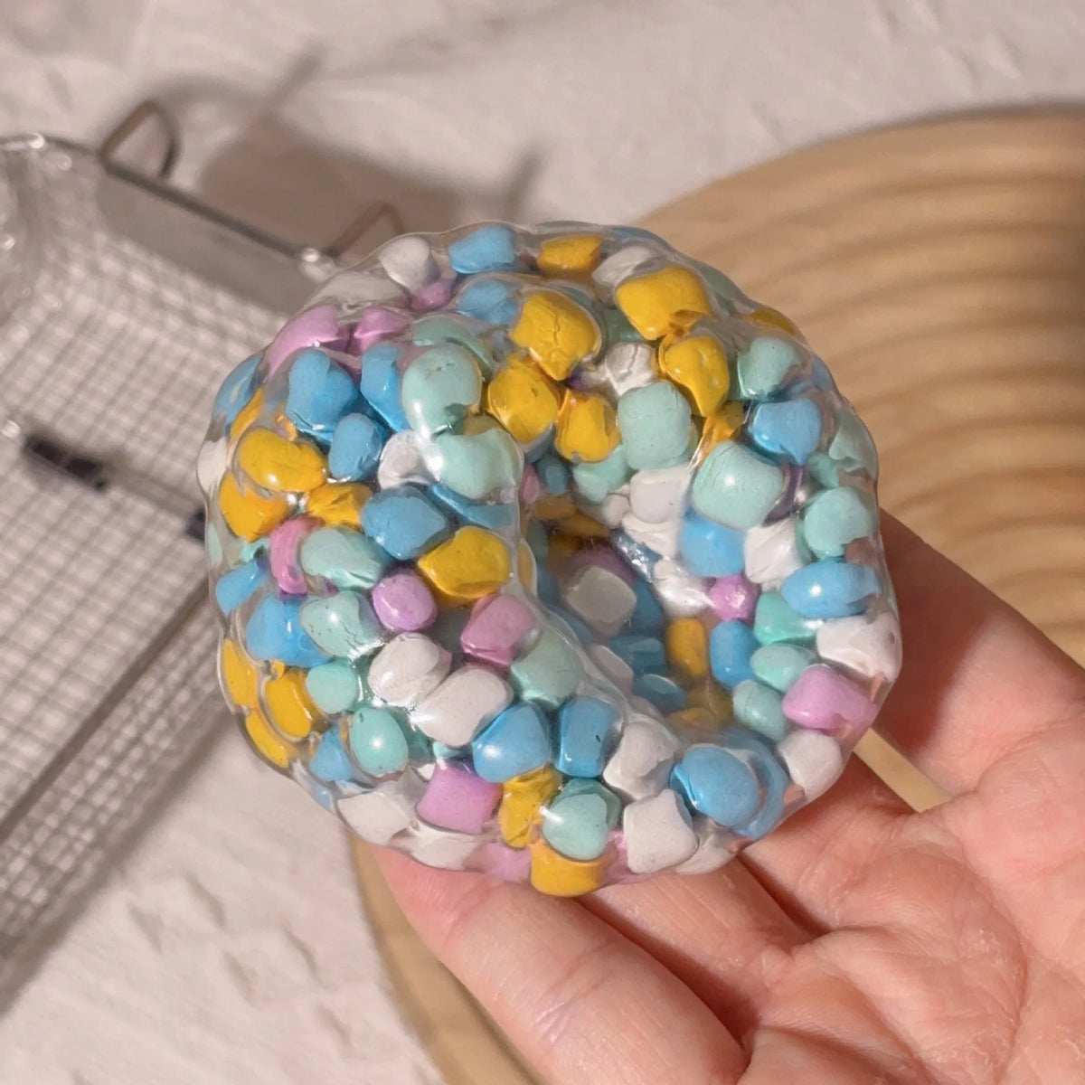 A hand holds a colorful squishy sensory stress ball filled with small, pastel-colored foam balls.