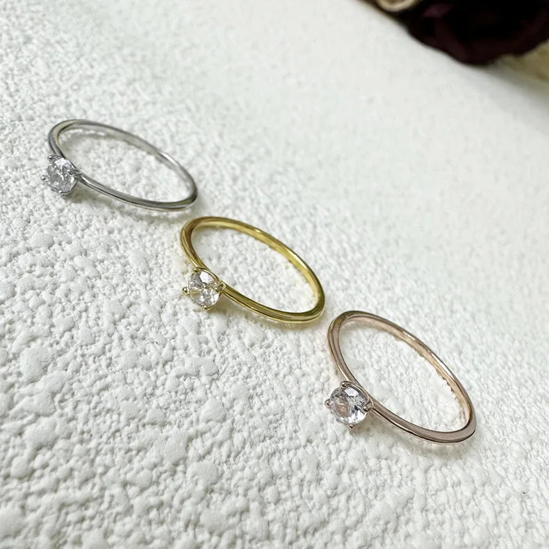 Three minimalist rings featuring clear solitaire stones in sterling silver and gold-toned metal on a white textured surface.
