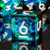 A close-up of a translucent blue resin polyhedral die showing the number 6, resting on a reflective surface.