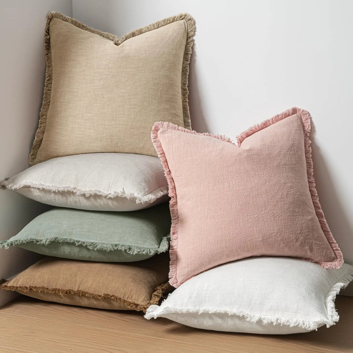 A stack of five boho fringe throw pillows in natural linen textures, including beige, pink, and sage green.