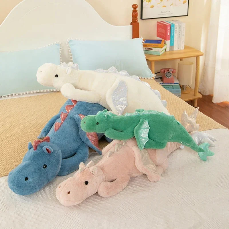 A collection of soft stuffed animal plush pillows featuring a white unicorn, blue dragon, and pink dinosaur resting on a bed.