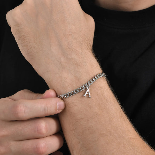 Primer plano de una mano vistiendo una pulsera de eslabones cubanos plateada con un dije inicial en la muñeca.
