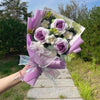 Handgemachter Blumenstrauß aus Häkelarbeit, Sonnenblumen, in lila Papier und Schleife, vor natürlichem Hintergrund.