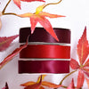 A stack of deep red and burgundy satin ribbon rolls against a white background with autumn leaves.