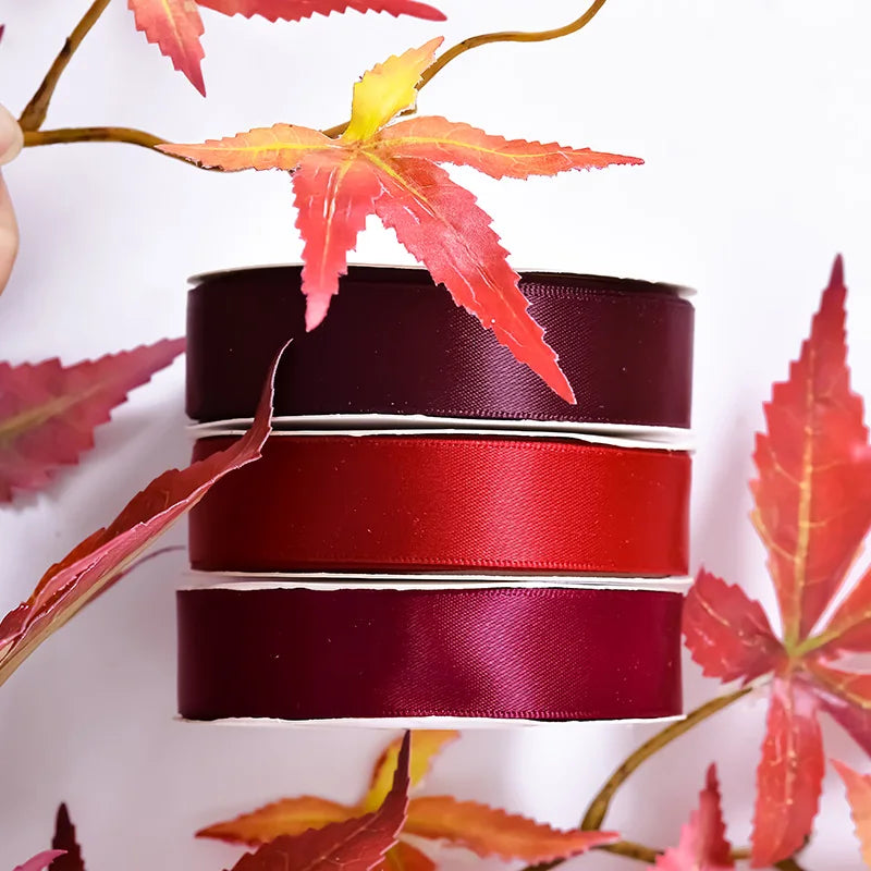 A stack of deep red and burgundy satin ribbon rolls against a white background with autumn leaves.