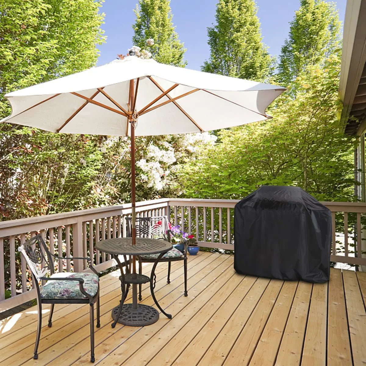 A black waterproof grill cover is placed on a wooden deck next to a table and chairs under a large white umbrella.