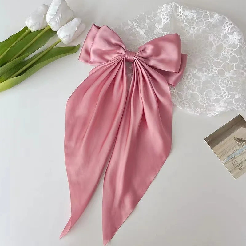 A pink silk bow hair clip with a ponytail holder is displayed next to white flowers and lace fabric.